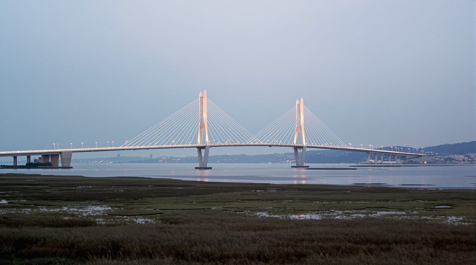 A cable-stayed bridge with two diamond-shaped towers over a wide river at dusk