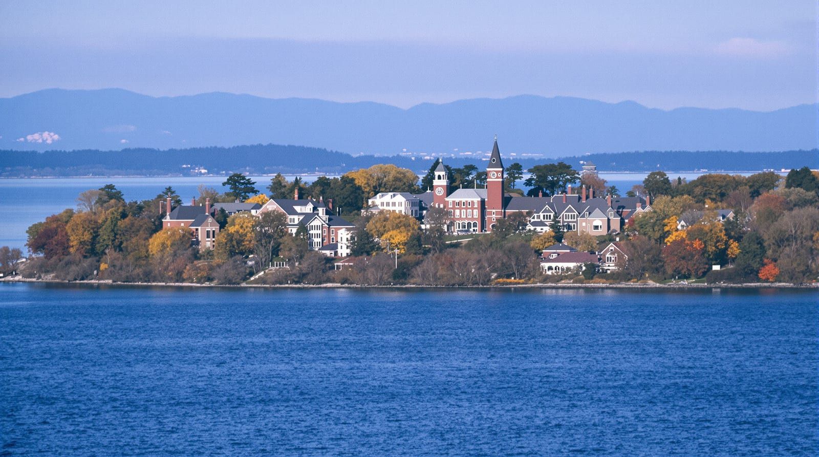 Red brick college buildings and a clock tower above a large lake with mountains behind