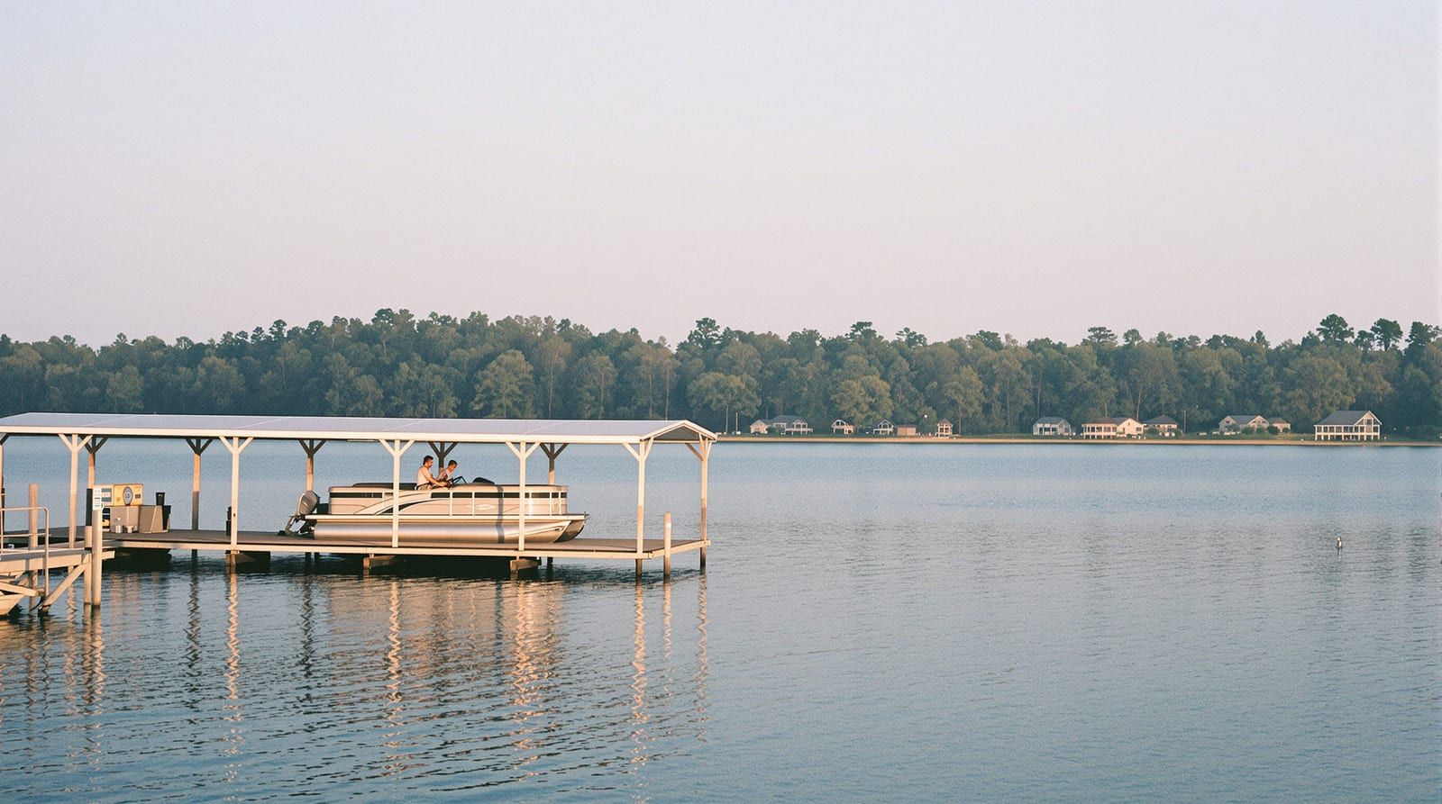 A covered boat dock with a pontoon boat on a wide lake at evening