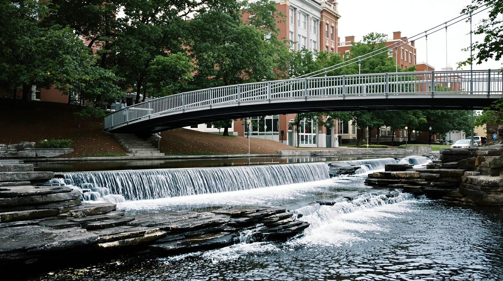 A curved pedestrian bridge over a small river falling across granite ledges in a downtown park